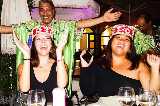 Two women sitting at a table, laughing and clapping, wearing colorful hats, with a smiling performer in a green outfit standing behind them.