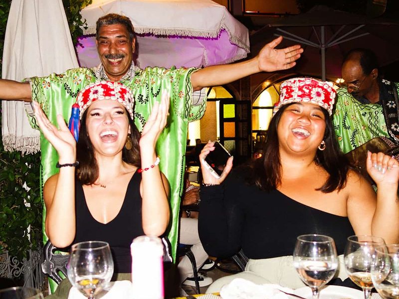 Two smiling women wearing festive hats clap at a table with drinks, while a man in a green shirt joyfully poses behind them.