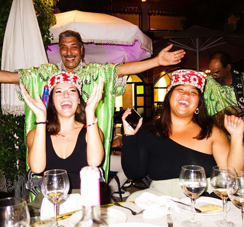 Two smiling women wearing festive hats clap at a table with drinks, while a man in a green shirt joyfully poses behind them.
