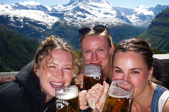 Three people smiling and holding beer mugs outdoors, with the Matterhorn mountain in the background under a clear blue sky.
