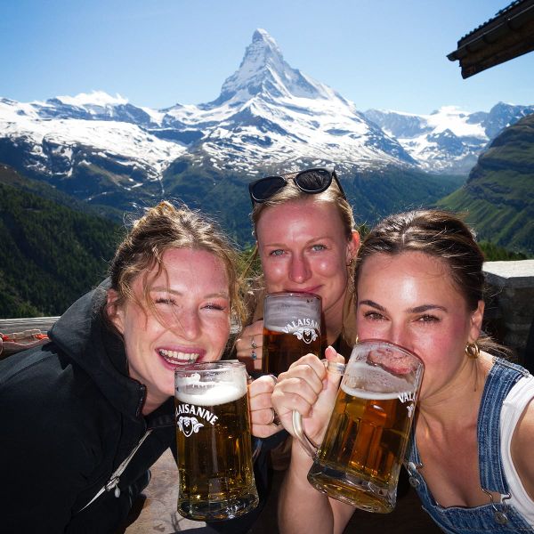 Three people smiling and holding beer mugs outdoors, with the Matterhorn mountain in the background under a clear blue sky.