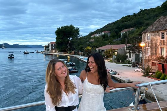 Two women smiling and enjoying a scenic waterfront view, sitting on a boat with a small village in the background under a cloudy sky.