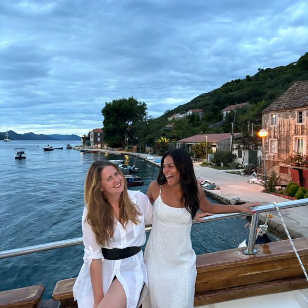 Two women smiling and enjoying a scenic waterfront view, sitting on a boat with a small village in the background under a cloudy sky.