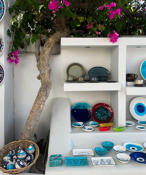 A display of colorful ceramic plates and bowls, including blue eye motifs, set against a white wall with pink flowers and a wicker basket.