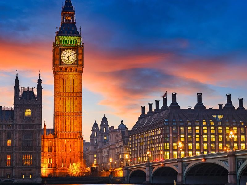 Big Ben at sunset with a colorful sky, reflected in the River Thames, and Westminster Bridge lit by streetlights.