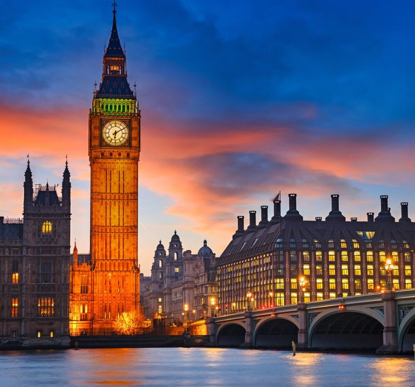 Big Ben at sunset with a colorful sky, reflected in the River Thames, and Westminster Bridge lit by streetlights.