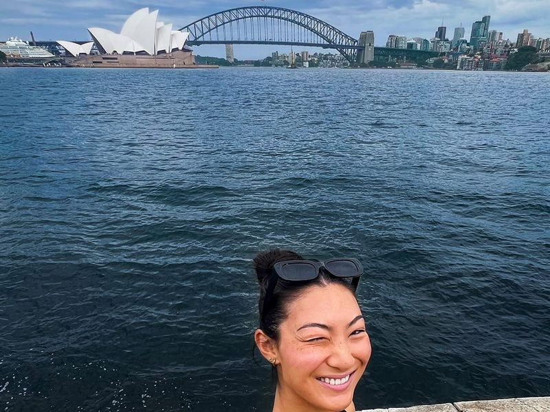 A person winks while taking a selfie near the water, with the Sydney Opera House and Harbour Bridge in the background under a cloudy sky.
