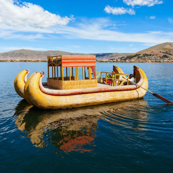 A traditional reed boat with a canopy floats on a lake under a bright blue sky, with distant hills and a shoreline in the background.