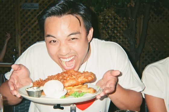 Person excitedly holding a plate of fish and chips, with mushy peas and tartar sauce, wearing a "I ♥ London" shirt at a wooden table.