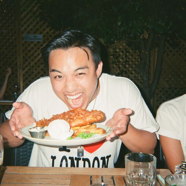 Person excitedly holding a plate of fish and chips, with mushy peas and tartar sauce, wearing a "I ♥ London" shirt at a wooden table.