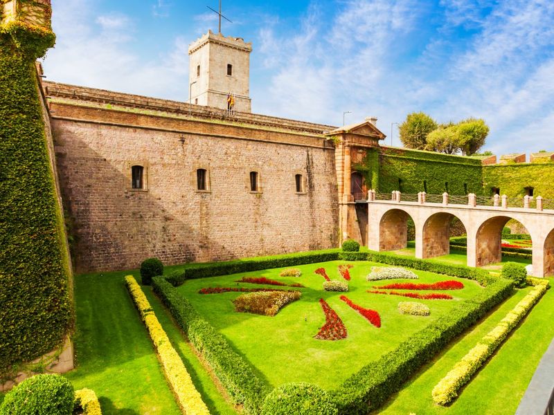 Stone fortress with a central tower and arched walkway, surrounded by manicured gardens featuring geometric patterns and lush greenery under a blue sky.