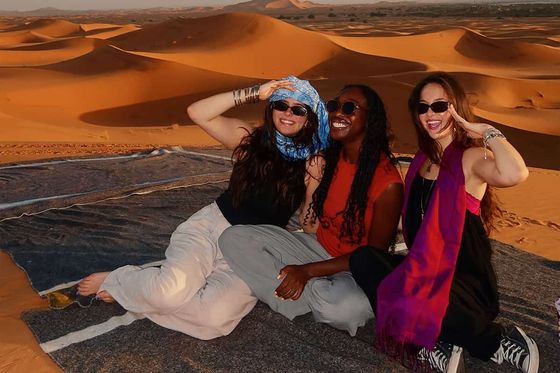 Three people sitting on a blanket in a desert, smiling and posing with dunes in the background under a partly cloudy sky.
