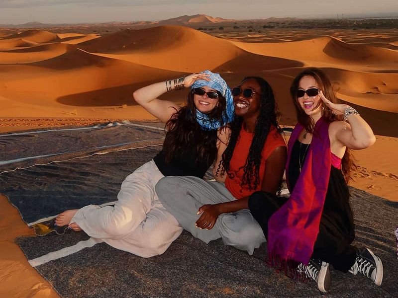 Three people sit on a blanket in a desert, smiling and posing with sand dunes in the background under a partly cloudy sky.