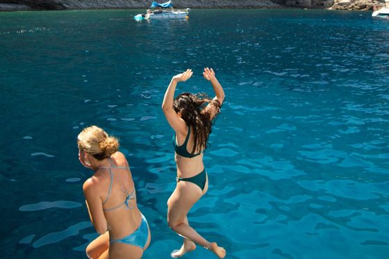 Two women in swimsuits jump into clear blue water near a rocky shoreline, with a sailboat in the background.