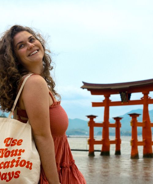 A smiling woman with curly hair holds a tote bag reading "Use Your Vacation Days" near a traditional red torii gate by the water.