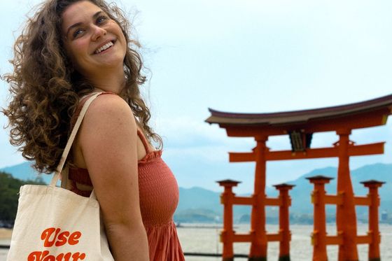 A smiling woman with curly hair holds a tote bag reading "Use Your Vacation Days" near a traditional red torii gate by the water.