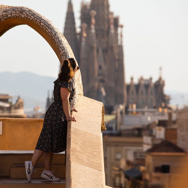 Woman in a floral dress leans on a rooftop railing, overlooking the cityscape with a prominent cathedral in the background.