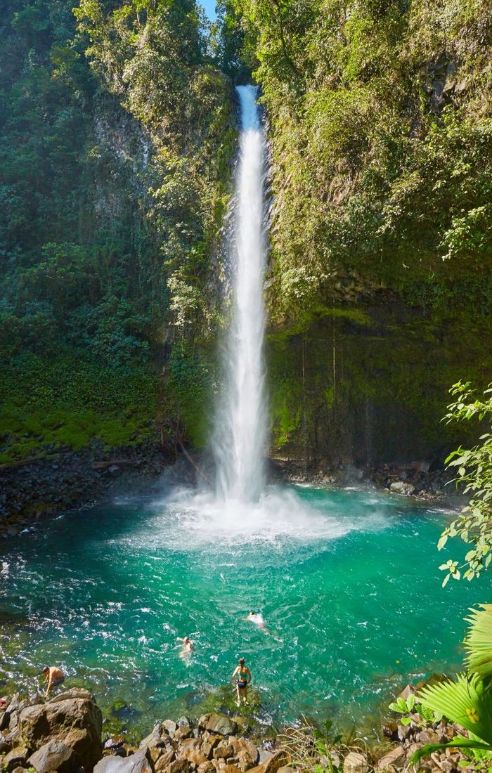 A tall waterfall cascading into a turquoise pool surrounded by lush greenery, with swimmers enjoying the water.