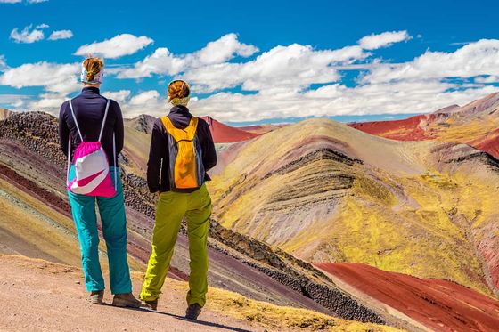 Two people in colorful outfits stand on a path, overlooking vibrant striped mountains under a bright blue sky with scattered clouds.
