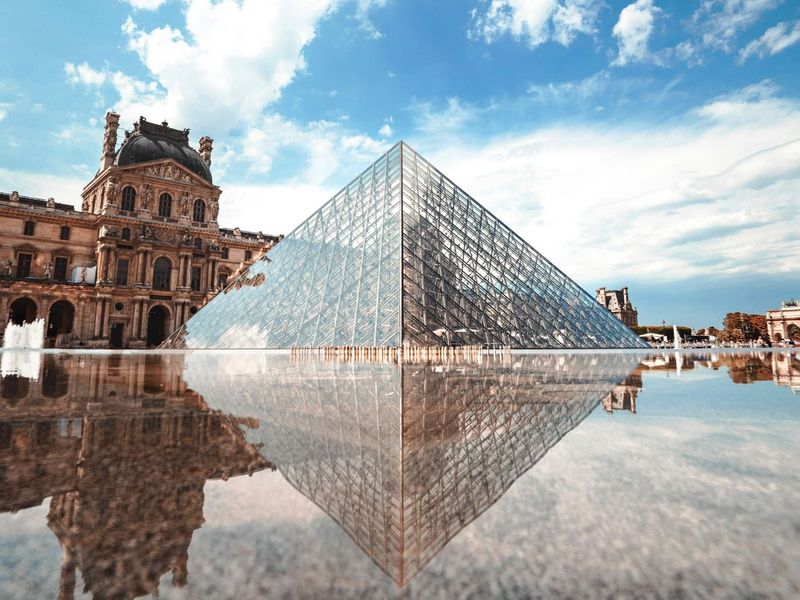 The Louvre Pyramid reflecting on a smooth water surface, with the historic museum building and a bright blue sky in the background.
