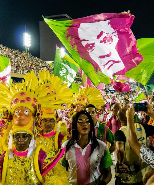 Carnival parade with vibrant costumes and large pink and green flags featuring a face, surrounded by a lively crowd at night.