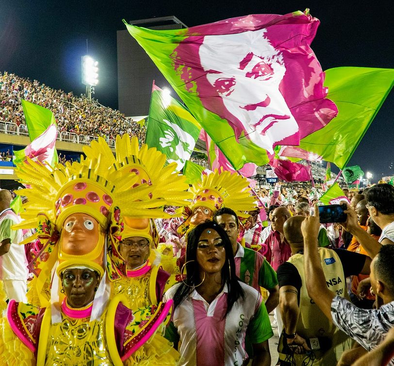 Carnival parade with vibrant costumes and large pink and green flags featuring a face, surrounded by a lively crowd at night.