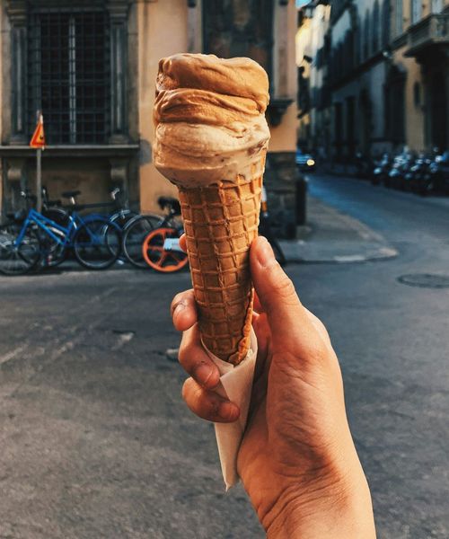 Hand holding a waffle cone with brown gelato, set against an urban street with parked bicycles and surrounding buildings in the background.