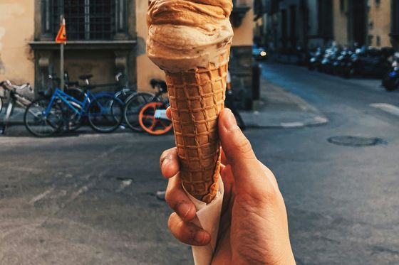 Hand holding a waffle cone with brown gelato, set against an urban street with parked bicycles and surrounding buildings in the background.