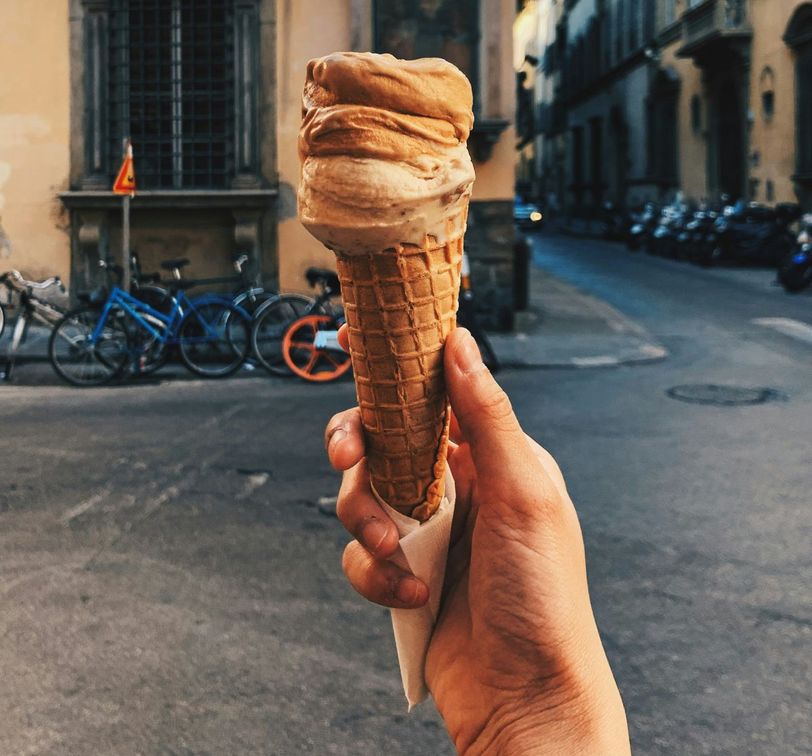 Hand holding a waffle cone with brown gelato, set against an urban street with parked bicycles and surrounding buildings in the background.