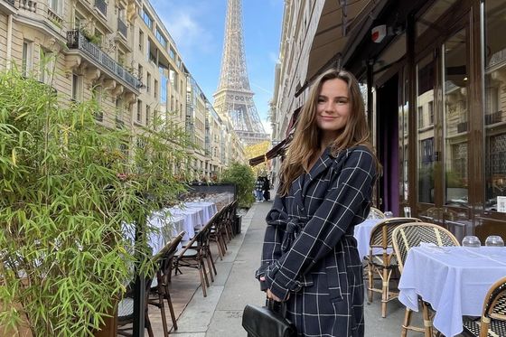 A woman in a checkered coat stands in a Parisian street with outdoor dining tables, the Eiffel Tower visible in the background.