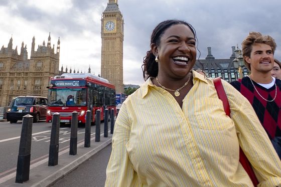 A smiling woman in a yellow shirt walks near Big Ben in London, with a red double-decker bus and cloudy sky in the background.