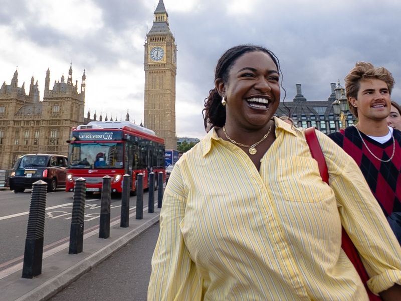 A smiling woman in a yellow shirt walks near Big Ben in London, with a red double-decker bus and cloudy sky in the background.