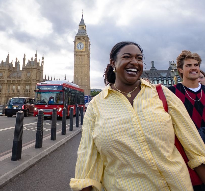 A smiling woman in a yellow shirt walks near Big Ben in London, with a red double-decker bus and cloudy sky in the background.