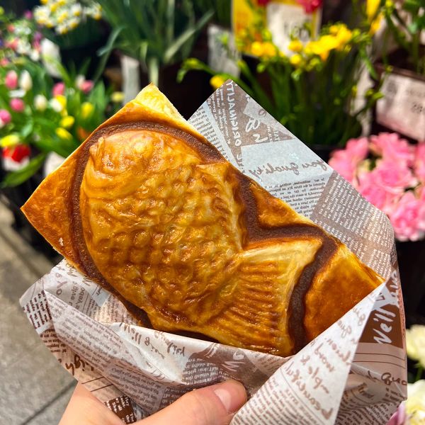 A hand holds a fish-shaped pastry wrapped in printed paper, with colorful flowers in the background.