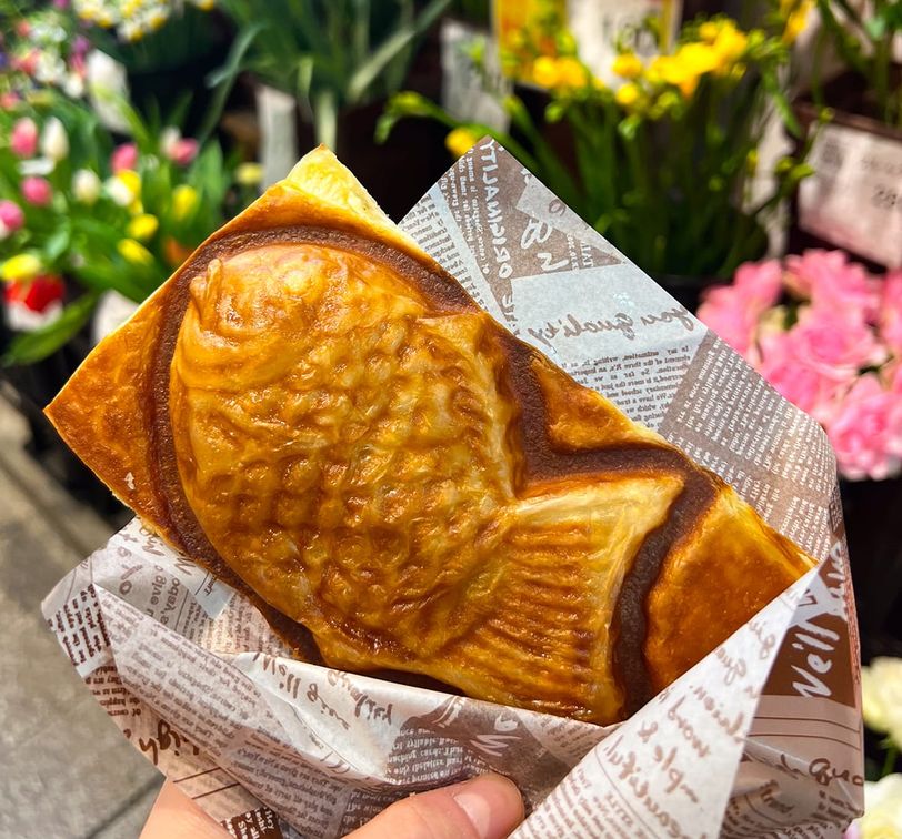 A hand holds a fish-shaped pastry wrapped in printed paper, with colorful flowers in the background.