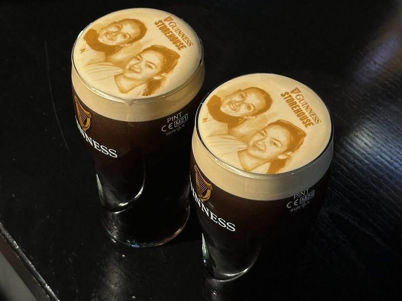 Two pints of Guinness with printed latte art images on the foam, set on a dark table.