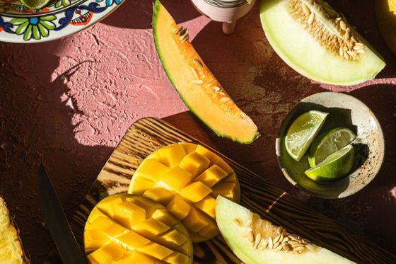 Assorted sliced fruits including mango, cantaloupe, honeydew, and lime on a decorative plate and wooden board, with a small cactus nearby.