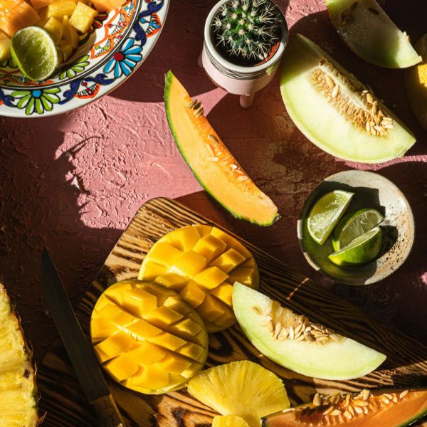 Assorted sliced fruits including mango, cantaloupe, honeydew, and lime on a decorative plate and wooden board, with a small cactus nearby.