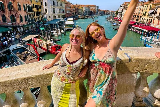Two women smiling and posing on a bridge in Venice with colorful buildings and boats along the canal in the background.
