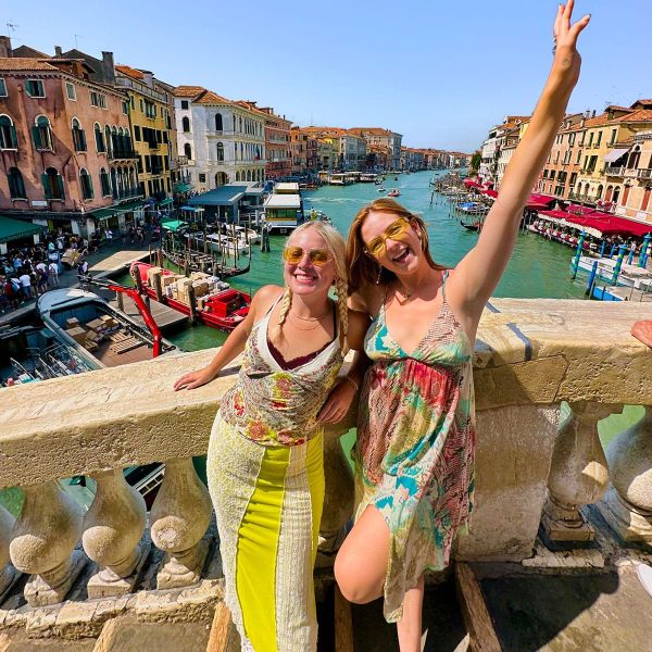 Two women smiling and posing on a bridge in Venice with colorful buildings and boats along the canal in the background.