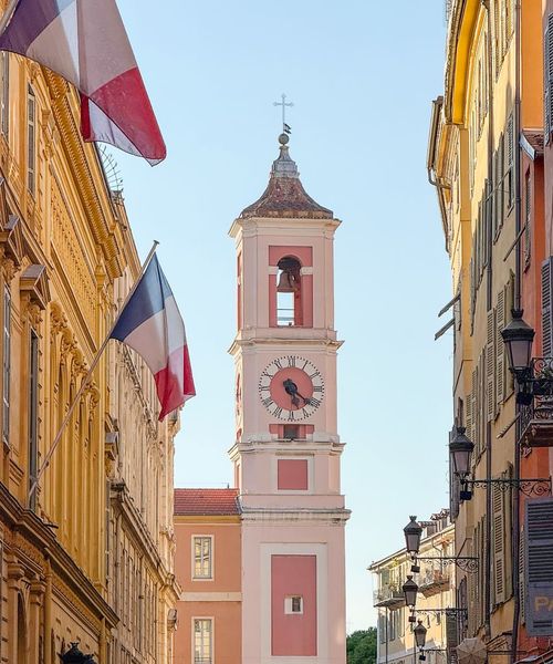 Clock tower between colorful buildings with French flags, under a clear blue sky.