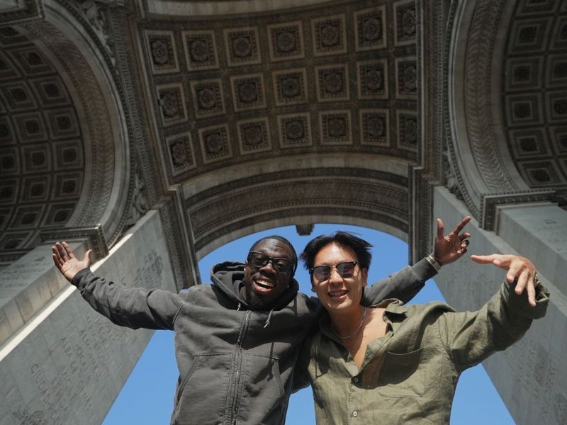 Two smiling friends with outstretched arms stand under the Arc de Triomphe, showcasing its intricate details against a clear blue sky.