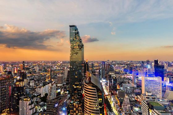 City skyline at dusk with a uniquely structured tall glass building, vibrant lights, and a colorful sky transitioning from orange to blue.