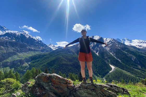 Person standing on a rocky hill with arms outstretched, surrounded by mountains and a bright sun above.