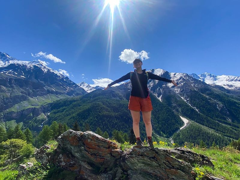 Person standing on a rocky hill with arms outstretched, surrounded by mountains and a bright sun above.