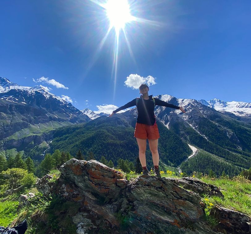 Person standing on a rocky hill with arms outstretched, surrounded by mountains and a bright sun above.