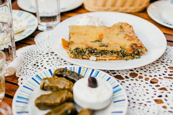 Spinach pie on a plate with a side of tzatziki, stuffed grape leaves, and a decorative table setting with glasses of water.
