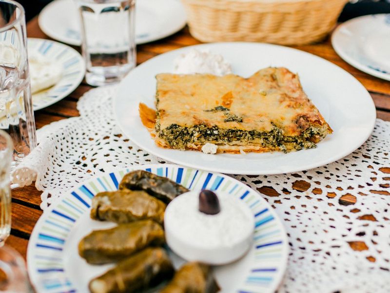 Spinach pie on a plate with a side of tzatziki, stuffed grape leaves, and a decorative table setting with glasses of water.