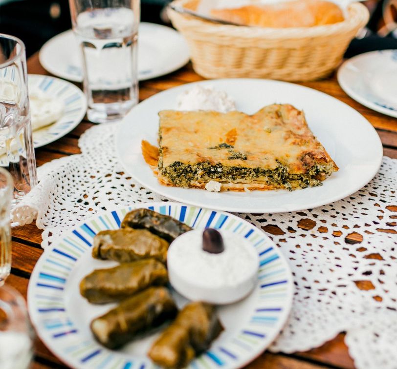 Spinach pie on a plate with a side of tzatziki, stuffed grape leaves, and a decorative table setting with glasses of water.