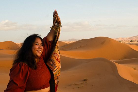 A person joyfully raises a colorful scarf in a sunlit desert landscape with rolling sand dunes under a clear blue sky.